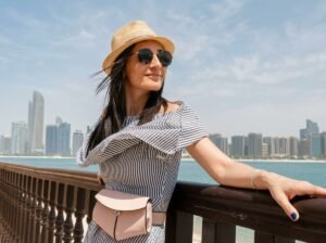A woman in a hat posing at a railing with the city skyline and waterfront behind her